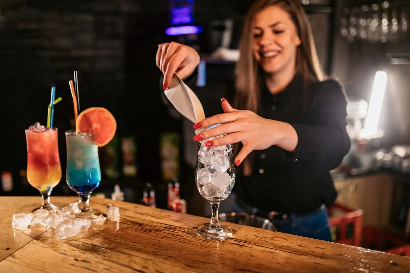 Bartender pouring ice into cocktail glass with vibrant blue and orange layered drinks on wooden bar counter