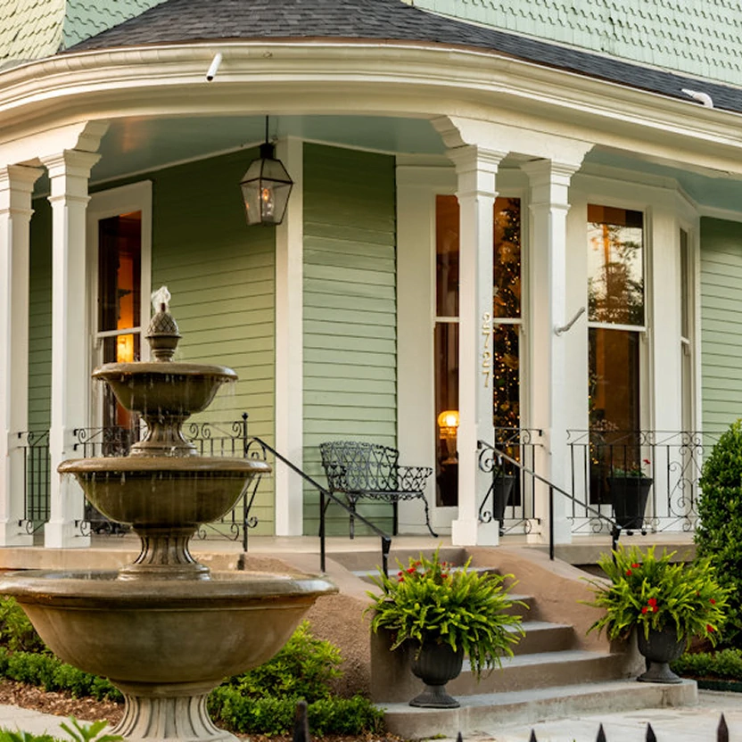  Elegant three-tiered fountain outside mint green Victorian home with white columns, black iron railing and potted plants 