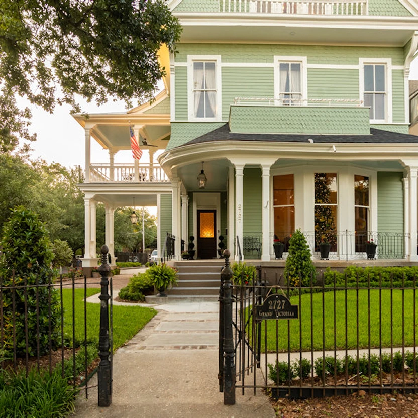  Mint green Victorian mansion with white columns, wraparound porch, manicured lawn and decorative black iron fence entrance 