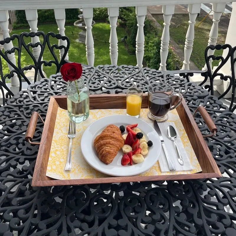  Elegant breakfast tray with croissant, fresh fruit, coffee, and orange juice on wrought iron patio furniture with garden view 