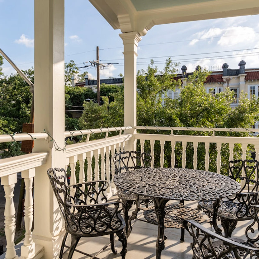  Covered porch with ornate white columns, black wrought iron table and chairs overlooking tree-lined New Orleans street 